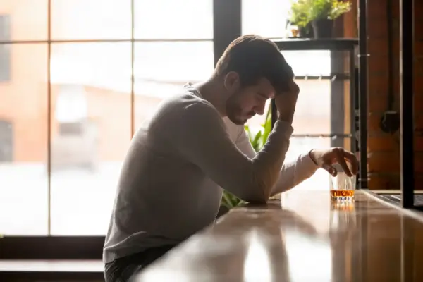 Despondent looking man sits at a bar with hand on his head and a glass of scotch