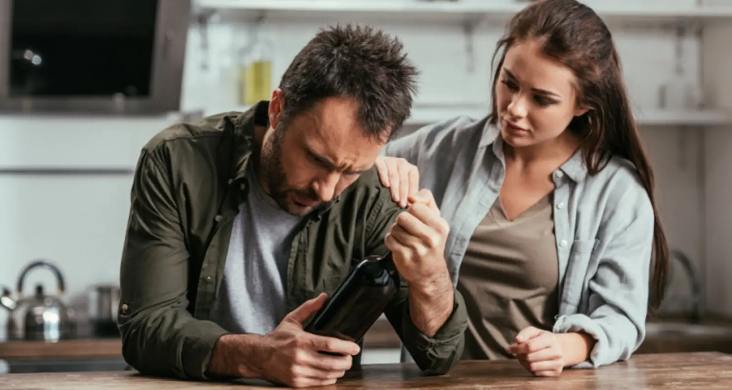 Sick-looking man holds a bottle of beer with concerned wife standing by